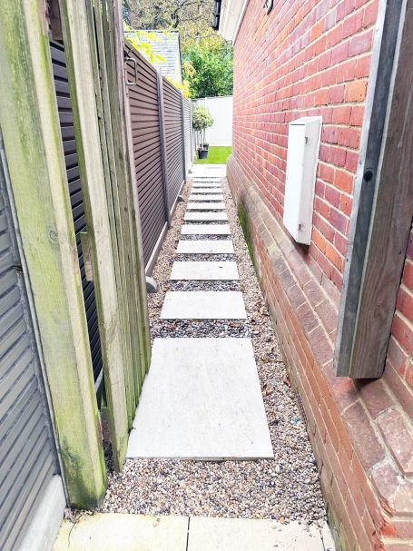 Narrow pathway between two buildings with concrete slabs and gravel.